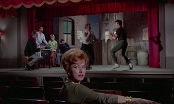 Movie still from “The Oscar” (1966), directed by Russell Rouse – A woman sitting in front of other people on a stage; Wide shot, Over the shoulder angle