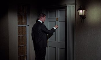 Movie still from “The Oscar” (1966), directed by Russell Rouse – A man in a black suit and white shirt standing in front of a white door; Medium shot, Low angle