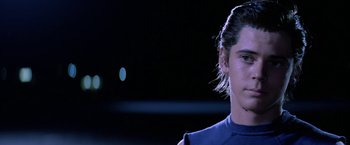 Movie still from “The Outsiders” (1983), directed by Francis Ford Coppola – A man with long dark hair standing in front of a black background; Close Up shot, Low angle