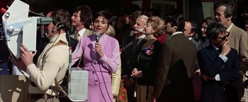 Movie still from “The Parallax View” (1974), directed by Alan J. Pakula – A group of people standing in a crowd and one woman is speaking into a microphone; Medium shot, Low angle