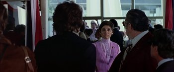 Movie still from “The Parallax View” (1974), directed by Alan J. Pakula – A woman in a purple dress is standing next to a man; Medium shot, Over the shoulder angle