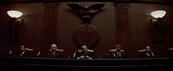 Movie still from “The Parallax View” (1974), directed by Alan J. Pakula – Three men in suits sitting in front of a court room sign; Wide shot, Low angle