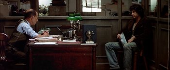 Movie still from “The Parallax View” (1974), directed by Alan J. Pakula – A man sitting at a desk in front of a green lamp; Wide shot, High angle