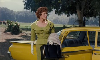 Movie still from “The Parent Trap” (1961), directed by David Swift – A woman in a green dress holding a black suitcase next to a yellow car; Medium shot, Low angle