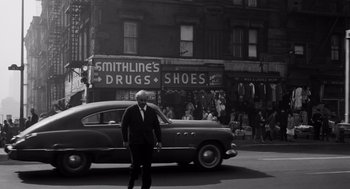 Movie still from “The Pawnbroker” (1964), directed by Sidney Lumet – A man in a suit and tie standing next to an old car; Wide shot, High angle
