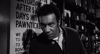 Movie still from “The Pawnbroker” (1964), directed by Sidney Lumet – A black - and - white photo of a man in front of signs; Close Up shot, Low angle