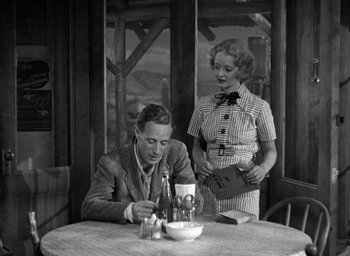 Movie still from “The Petrified Forest” (1936), directed by Archie Mayo – A man sitting at a table with a bottle of wine in front of a woman standing next to him; Medium shot, High angle