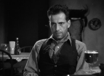 Movie still from “The Petrified Forest” (1936), directed by Archie Mayo – A black and white photo of a man sitting at a table with a drink; Medium shot, Over the shoulder angle