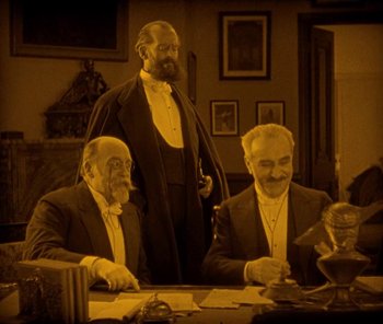Movie still from “The Phantom of the Opera” (1925), directed by Lon Chaney – A man standing next to two other men at a table; Medium shot, High angle