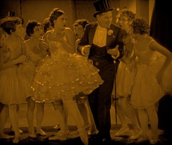 Movie still from “The Phantom of the Opera” (1925), directed by Lon Chaney – A man in a top hat is walking next to a group of women; Wide shot, High angle