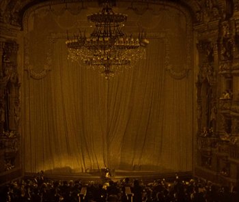 A large chandelier hangs in front of a stage with people sitting on it