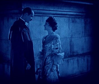 Movie still from “The Phantom of the Opera” (1925), directed by Lon Chaney – A man and a woman standing next to each other in front of a stone wall; Medium shot, High angle