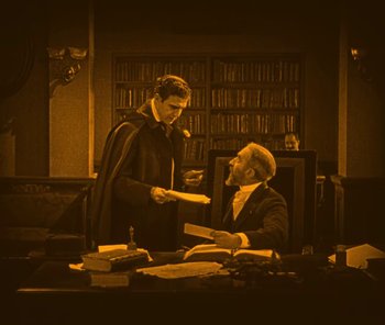 Movie still from “The Phantom of the Opera” (1925), directed by Lon Chaney – Two men are sitting at a desk and one is holding a book; Wide shot, High angle