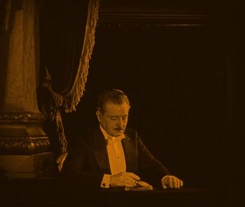 Movie still from “The Phantom of the Opera” (1925), directed by Lon Chaney – A man in a tuxedo sitting at a table in front of a curtain; Medium shot, High angle