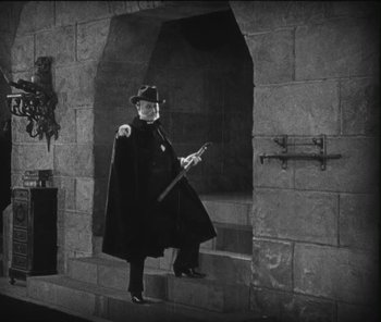 Movie still from “The Phantom of the Opera” (1925), directed by Lon Chaney – A black and white photo of a man in a black coat and hat; Wide shot, Low angle