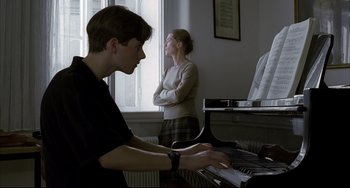 Movie still from “The Piano Teacher” (2001), directed by Michael Haneke – A young man playing the piano as a woman looks on; Medium shot, Low angle