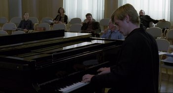 Movie still from “The Piano Teacher” (2001), directed by Michael Haneke – A man playing a piano in front of a group of people in a room; Medium shot, Over the shoulder angle