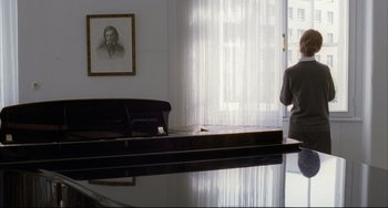 Movie still from “The Piano Teacher” (2001), directed by Michael Haneke – A man standing in front of a piano in a living room; Wide shot, Over the shoulder angle