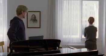 Movie still from “The Piano Teacher” (2001), directed by Michael Haneke – A man standing in front of a piano in a living room; Medium shot, Over the shoulder angle