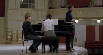 Movie still from “The Piano Teacher” (2001), directed by Michael Haneke – Three people are sitting at a piano in a room; Wide shot, Low angle