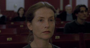 Movie still from “The Piano Teacher” (2001), directed by Michael Haneke – A person sitting in front of a group of people; Close Up shot, Over the shoulder angle