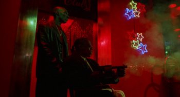 Movie still from “The Players Club” (1998), directed by Ice Cube – A man sitting next to another man holding a gun in a room; Wide shot, Low angle