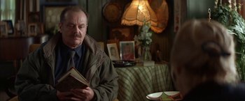 Movie still from “The Pledge” (2001), directed by Sean Penn – An older man sitting at a table holding a book; Medium shot, Over the shoulder angle