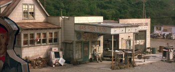 Movie still from “The Pledge” (2001), directed by Sean Penn – A man sitting on the side of the road in front of a gas station; Extreme Wide shot, High angle