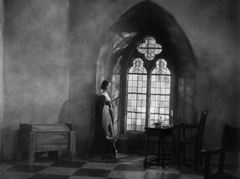 Movie still from “The Private Life of Henry VIII” (1933), directed by Alexander Korda – A black and white photo of a woman standing in front of a stained glass window; Wide shot, Low angle