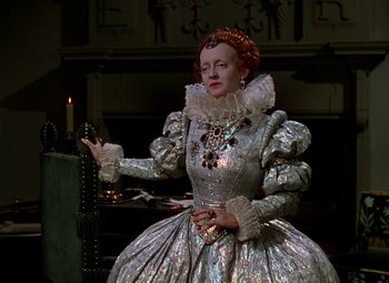 Movie still from “The Private Lives of Elizabeth and Essex” (1939), directed by Michael Curtiz – A woman dressed in a silver dress and a white wig; Medium shot, Over the shoulder angle