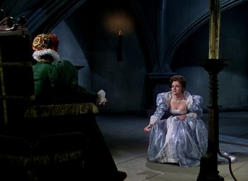 Movie still from “The Private Lives of Elizabeth and Essex” (1939), directed by Michael Curtiz – A woman dressed in a gown and a hat is sitting in front of a man; Wide shot, Over the shoulder angle