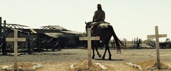Movie still from “The Proposition” (2005), directed by John Hillcoat – A man riding a horse in front of a cross on a dirt road; Wide shot, Low angle
