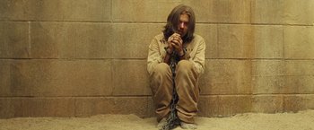 Movie still from “The Proposition” (2005), directed by John Hillcoat – A man sitting on the ground chained to a wall; Medium shot, High angle