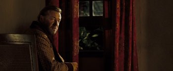 Movie still from “The Proposition” (2005), directed by John Hillcoat – A man standing in front of a window with red drapes; Medium shot, Over the shoulder angle