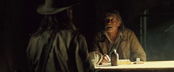 Movie still from “The Proposition” (2005), directed by John Hillcoat – An older man sitting at a table with a bottle; Medium shot, Over the shoulder angle