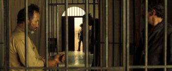 Movie still from “The Proposition” (2005), directed by John Hillcoat – A man standing in a jail cell behind bars; Wide shot, Over the shoulder angle