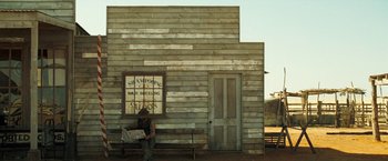 Movie still from “The Proposition” (2005), directed by John Hillcoat – A man sitting on a bench in front of a building; Wide shot, Low angle