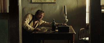 Movie still from “The Proposition” (2005), directed by John Hillcoat – A man sitting at a wooden table with a bottle of wine; Medium shot, Low angle