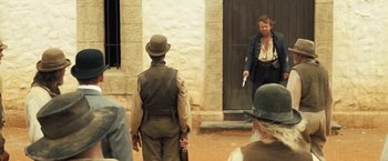 Movie still from “The Proposition” (2005), directed by John Hillcoat – A man holding a gun in front of another man in front of a building; Medium shot, Over the shoulder angle