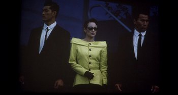 Movie still from “The Punisher” (1989), directed by Mark Goldblatt – A woman in a yellow jacket standing in front of two men in black suits; Medium shot, Low angle
