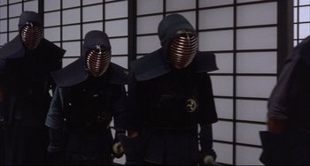 Movie still from “The Punisher” (1989), directed by Mark Goldblatt – Two people in black suits and helmets are standing in front of a wall; Medium shot, Low angle