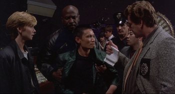 Movie still from “The Punisher” (1989), directed by Mark Goldblatt – A group of people standing around a man in a green jacket; Medium shot, Over the shoulder angle