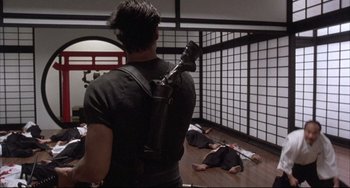 Movie still from “The Punisher” (1989), directed by Mark Goldblatt – A man with a gun strapped to his back is standing in front of a group of people laying on the floor; Wide shot, Over the shoulder angle
