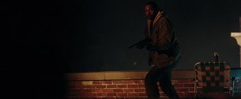 Movie still from “The Purge: Election Year” (2016), directed by James DeMonaco – A man holding a gun while standing next to a brick wall; Medium shot, Low angle