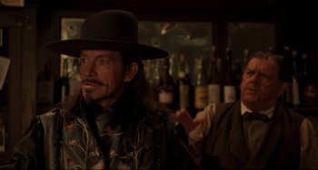 Movie still from “The Quick and the Dead” (1995), directed by Sam Raimi – A man with a hat and a mustache in front of a bar; Medium shot, Over the shoulder angle