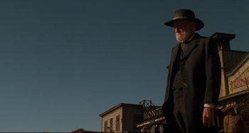 Movie still from “The Quick and the Dead” (1995), directed by Sam Raimi – An older man in a hat and a suit stands in front of a building; Medium shot, Low angle