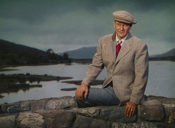 Movie still from “The Quiet Man” (1952), directed by John Ford – A man sitting on top of a stone wall near a body of water; Medium shot, Low angle