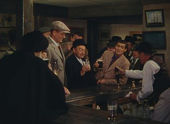 Movie still from “The Quiet Man” (1952), directed by John Ford – A group of men standing around a bar with drinks; Wide shot, High angle