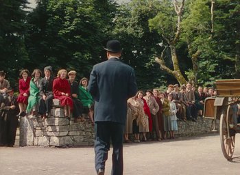 Movie still from “The Quiet Man” (1952), directed by John Ford – A crowd of people sitting on a stone wall watching a man walk; Wide shot, Over the shoulder angle