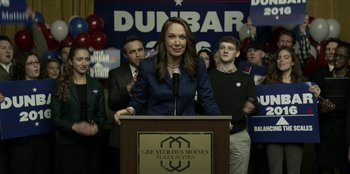 Movie still from “House of Cards” (2013), created by Beau Willimon – A woman standing at a podium in front of people; Medium shot, High angle